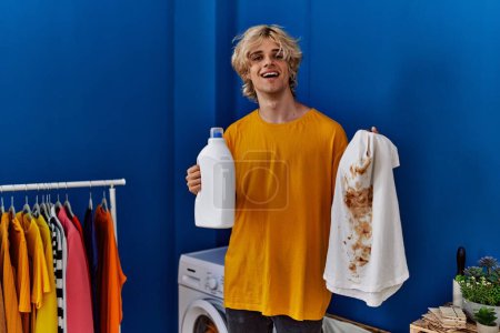 Photo for Young blond man holding dirty t shirt and detergent at laundry room - Royalty Free Image