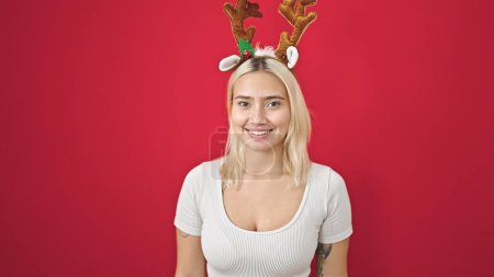 Foto de Joven hermosa mujer hispana sonriendo confiada usando sombrero de Navidad de renos sobre fondo rojo aislado - Imagen libre de derechos