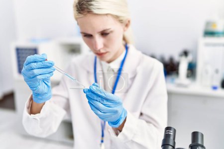 Foto de Joven mujer rubia científica vertiendo líquido en la muestra en el laboratorio - Imagen libre de derechos