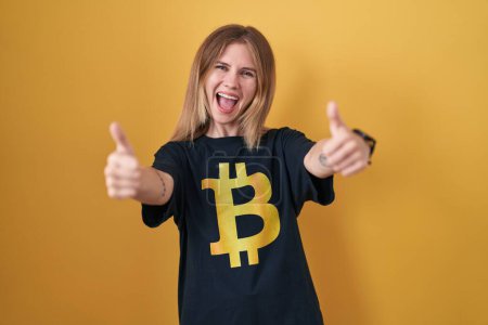 Foto de Mujer rubia caucásica con camiseta bitcoin aprobando hacer un gesto positivo con la mano, los pulgares hacia arriba sonriendo y feliz por el éxito. gesto ganador. - Imagen libre de derechos