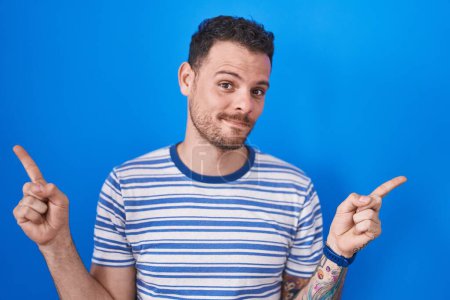 Téléchargez les photos : Jeune homme hispanique debout sur fond bleu souriant confiant pointant avec les doigts vers différentes directions. espace de copie pour la publicité - en image libre de droit