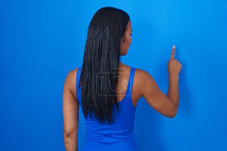 Photo for Hispanic woman standing over blue background posing backwards pointing ahead with finger hand - Royalty Free Image
