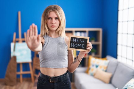 Photo for Blonde caucasian woman holding blackboard with new home text with open hand doing stop sign with serious and confident expression, defense gesture - Royalty Free Image