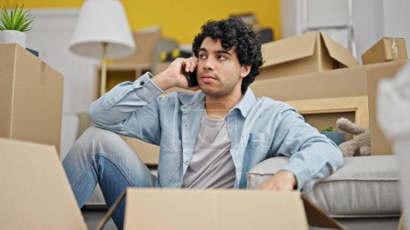Photo for Young latin man talking on smartphone unpacking cardboard box looking upset at new home - Royalty Free Image