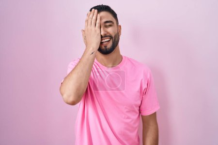 Téléchargez les photos : Jeune homme hispanique debout sur fond rose couvrant un oeil avec la main, sourire confiant sur le visage et l'émotion surprise. - en image libre de droit