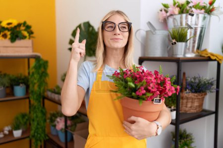 Foto de Mujer joven caucásica que trabaja en floristería sosteniendo la planta sonriendo con una idea o pregunta señalando el dedo con la cara feliz, número uno - Imagen libre de derechos