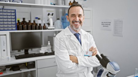 Téléchargez les photos : Un scientifique souriant d'âge moyen aux cheveux gris croise les bras, travaille au microscope dans son laboratoire - en image libre de droit