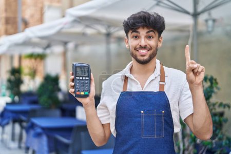 Téléchargez les photos : Homme arabe avec barbe portant tablier de serveur à la terrasse du restaurant tenant dataphone souriant avec une idée ou une question pointant du doigt avec un visage heureux, numéro un - en image libre de droit