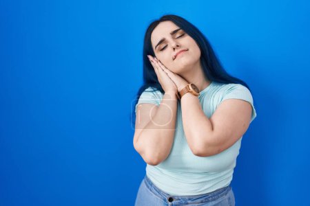 Photo for Young modern girl with blue hair standing over blue background sleeping tired dreaming and posing with hands together while smiling with closed eyes. - Royalty Free Image