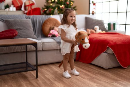 Foto de Adorable hispanic girl playing with horse toy standing by christmas tree at home - Imagen libre de derechos