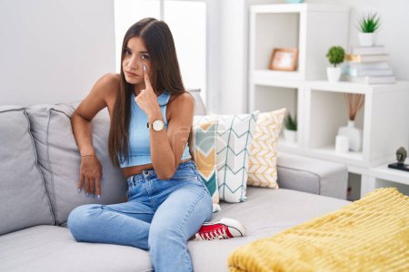 Foto de Young brunette woman sitting on the sofa at home pointing to the eye watching you gesture, suspicious expression - Imagen libre de derechos