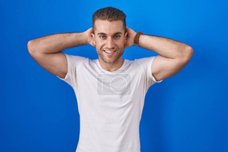 Téléchargez les photos : Jeune homme caucasien debout sur fond bleu relaxant et étirant, bras et mains derrière la tête et le cou souriant heureux - en image libre de droit