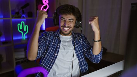 Foto de Hombre árabe joven guapo, un jugador seguro de sí mismo, transmite victoria nocturna en su videojuego, animando en la sala de juegos, sonrisa victoriosa iluminada por el resplandor de la pantalla. - Imagen libre de derechos