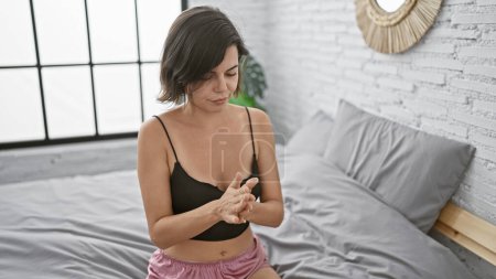 Téléchargez les photos : Belle jeune femme hispanique en pyjama appliquant un traitement de la peau, reposant sur un lit confortable dans une routine de relaxation matinale dans sa chambre à coucher à la maison - en image libre de droit