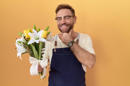 Foto de Hombre de mediana edad con floristería barba sosteniendo flores mirando con confianza a la cámara sonriendo con los brazos cruzados y la mano levantada en la barbilla. pensar positivamente. - Imagen libre de derechos