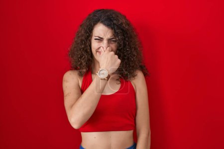 Téléchargez les photos : Femme hispanique avec les cheveux bouclés debout sur fond rouge sentant quelque chose de puant et dégoûtant, odeur intolérable, retenant souffle avec les doigts sur le nez. mauvaise odeur - en image libre de droit