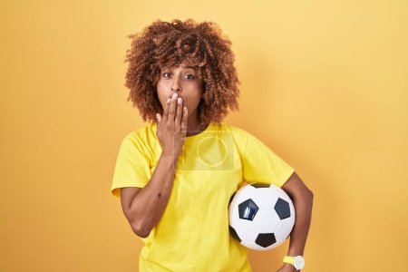 Téléchargez les photos : Jeune femme hispanique aux cheveux bouclés tenant ballon de football couvrant la bouche avec la main, choquée et effrayée par l'erreur. expression surprise - en image libre de droit