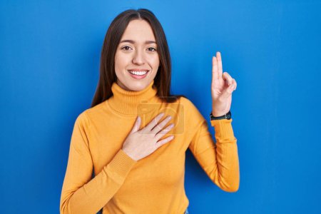 Photo for Young brunette woman standing over blue background smiling swearing with hand on chest and fingers up, making a loyalty promise oath - Royalty Free Image