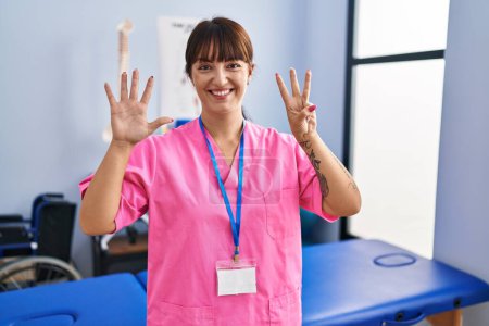 Foto de Joven morena que trabaja en la clínica de rehabilitación mostrando y señalando con los dedos número ocho mientras sonríe confiada y feliz. - Imagen libre de derechos