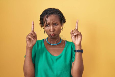 Téléchargez les photos : Femme africaine avec dreadlocks debout sur fond jaune pointant vers le haut regardant triste et contrarié, indiquant la direction avec les doigts, malheureux et déprimé. - en image libre de droit