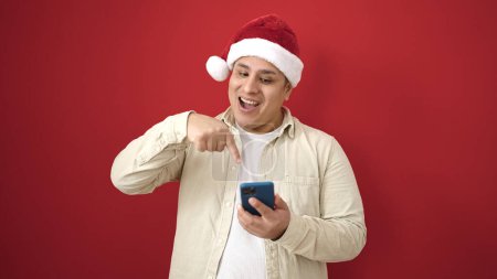 Téléchargez les photos : Jeune homme hispanique portant un chapeau de Noël pointant vers le smartphone sur fond rouge isolé - en image libre de droit