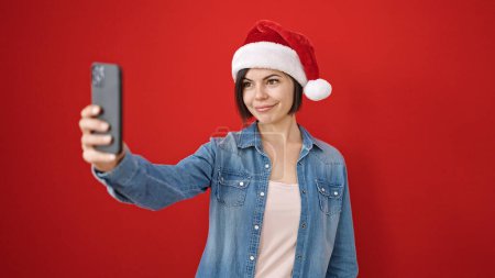 Foto de Joven mujer caucásica tomando una foto selfie usando sombrero de Navidad sobre fondo rojo aislado - Imagen libre de derechos
