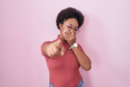 Téléchargez les photos : Belle femme africaine aux cheveux bouclés debout sur fond rose se moquant de vous, pointant du doigt l'appareil photo avec la main sur la bouche, expression de honte - en image libre de droit