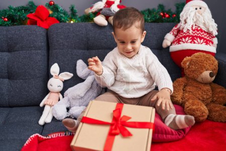 Foto de Adorable niño hispano desempacando regalo de Navidad sentado en un sofá en casa - Imagen libre de derechos