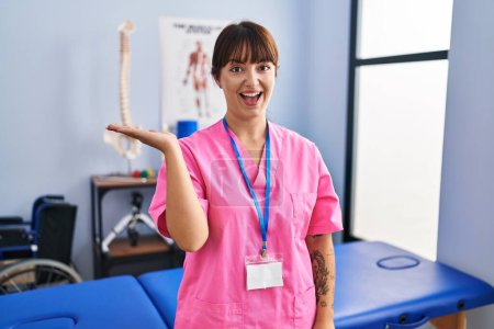 Photo for Young brunette woman working at rehabilitation clinic smiling cheerful presenting and pointing with palm of hand looking at the camera. - Royalty Free Image