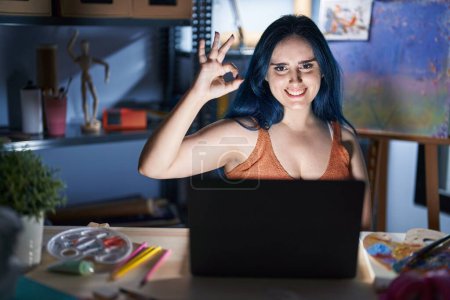 Téléchargez les photos : Jeune fille moderne avec les cheveux bleus assis au studio d'art avec ordinateur portable la nuit souriant positif faire ok signe avec la main et les doigts. expression réussie. - en image libre de droit