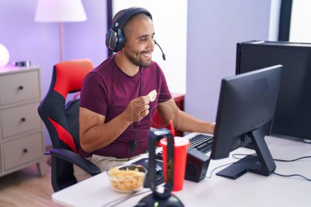 Photo for Young latin man streamer playing video game eating chips potatoes at gaming room - Royalty Free Image