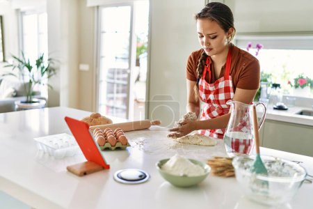 Photo for Young beautiful hispanic woman smiling confident kneading bread dough watching online recipe on touchpad at the kitchen - Royalty Free Image