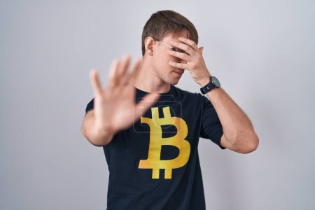 Téléchargez les photos : Homme blond caucasien portant Bitcoin t-shirt couvrant les yeux avec les mains et faire arrêter geste avec expression triste et peur. concept embarrassé et négatif. - en image libre de droit