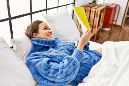 Foto de Joven hermosa mujer hispana leyendo libro acostado en la cama en el dormitorio - Imagen libre de derechos