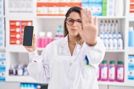 Photo for Blonde woman working at pharmacy drugstore showing smartphone screen with open hand doing stop sign with serious and confident expression, defense gesture - Royalty Free Image