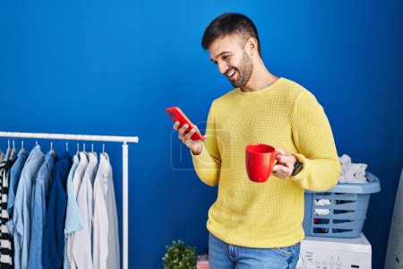 Photo for Young hispanic man using smartphone drinking coffee waiting for washing machine at laundry room - Royalty Free Image