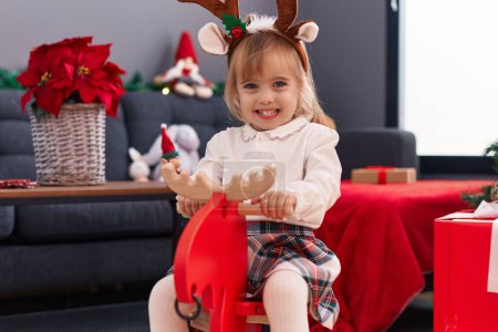 Photo for Adorable blonde girl playing with reindeer rocking by christmas tree at home - Royalty Free Image