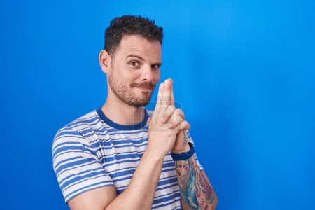 Photo for Young hispanic man standing over blue background holding symbolic gun with hand gesture, playing killing shooting weapons, angry face - Royalty Free Image