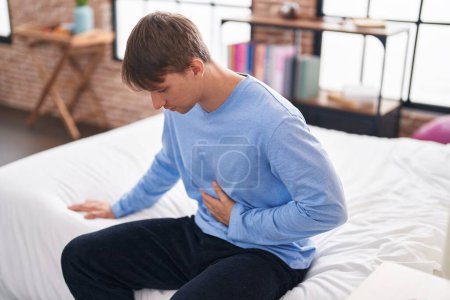 Téléchargez les photos : Jeune homme caucasien souffrant de maux d'estomac assis sur le lit dans la chambre - en image libre de droit