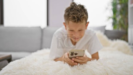 Téléchargez les photos : Adorable garçon blond, plein de joie, utilisant joyeusement son smartphone, envoyant des SMS alors qu'il était allongé sur un canapé, à l'intérieur, à la maison - en image libre de droit