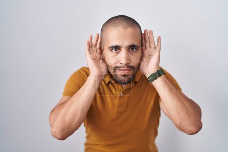 Téléchargez les photos : Hispanic man with beard standing over white background trying to hear both hands on ear gesture, curious for gossip. hearing problem, deaf - en image libre de droit