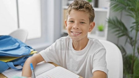 Téléchargez les photos : Joyeux garçon blond étudiant absorbé dans la prise de notes à son bureau, souriant dans l'ambiance de classe accueillante, montrant l'enthousiasme académique. - en image libre de droit