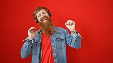Foto de Confiado joven pelirrojo amante de su música, bailando y divirtiéndose, sobre un fondo rojo aislado mientras escucha en atuendo casual, supurando positividad y felicidad - Imagen libre de derechos