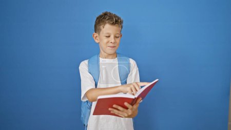 Photo for Adorable little blond boy student confidently smiling, joyfully reading a book while standing against an isolated blue background. - Royalty Free Image