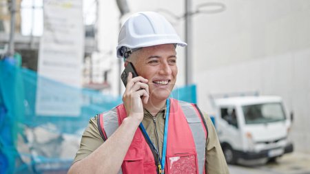 Foto de Joven hombre caucásico constructor sonriendo confiado hablando en el teléfono inteligente en el lugar de construcción - Imagen libre de derechos