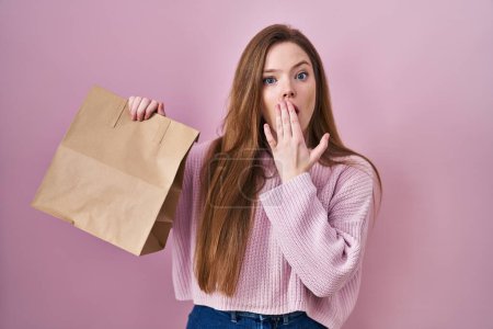 Foto de Joven mujer caucásica sosteniendo bolsa de la compra y tarjeta de crédito que cubre la boca con la mano, conmocionada y temerosa de error. expresión sorprendida - Imagen libre de derechos