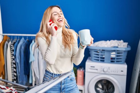 Téléchargez les photos : Young blonde woman talking on smartphone drinking coffee waiting for washing machine at laundry room - en image libre de droit