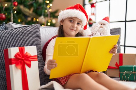 Photo for Adorable hispanic girl reading book sitting on sofa by christmas tree at home - Royalty Free Image