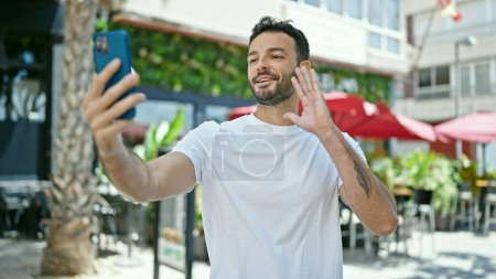 Téléchargez les photos : Jeune homme hispanique souriant confiant ayant appel vidéo à la terrasse du café - en image libre de droit
