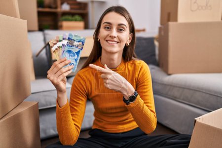 Téléchargez les photos : Jeune femme caucasienne assise sur le sol à la nouvelle maison tenant de l'argent canadien souriant heureux pointant avec la main et le doigt - en image libre de droit